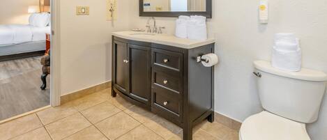 A hotel bathroom with a white toilet, a dark wooden vanity with drawers, a mirror above the sink, and a bed visible in the adjacent room.