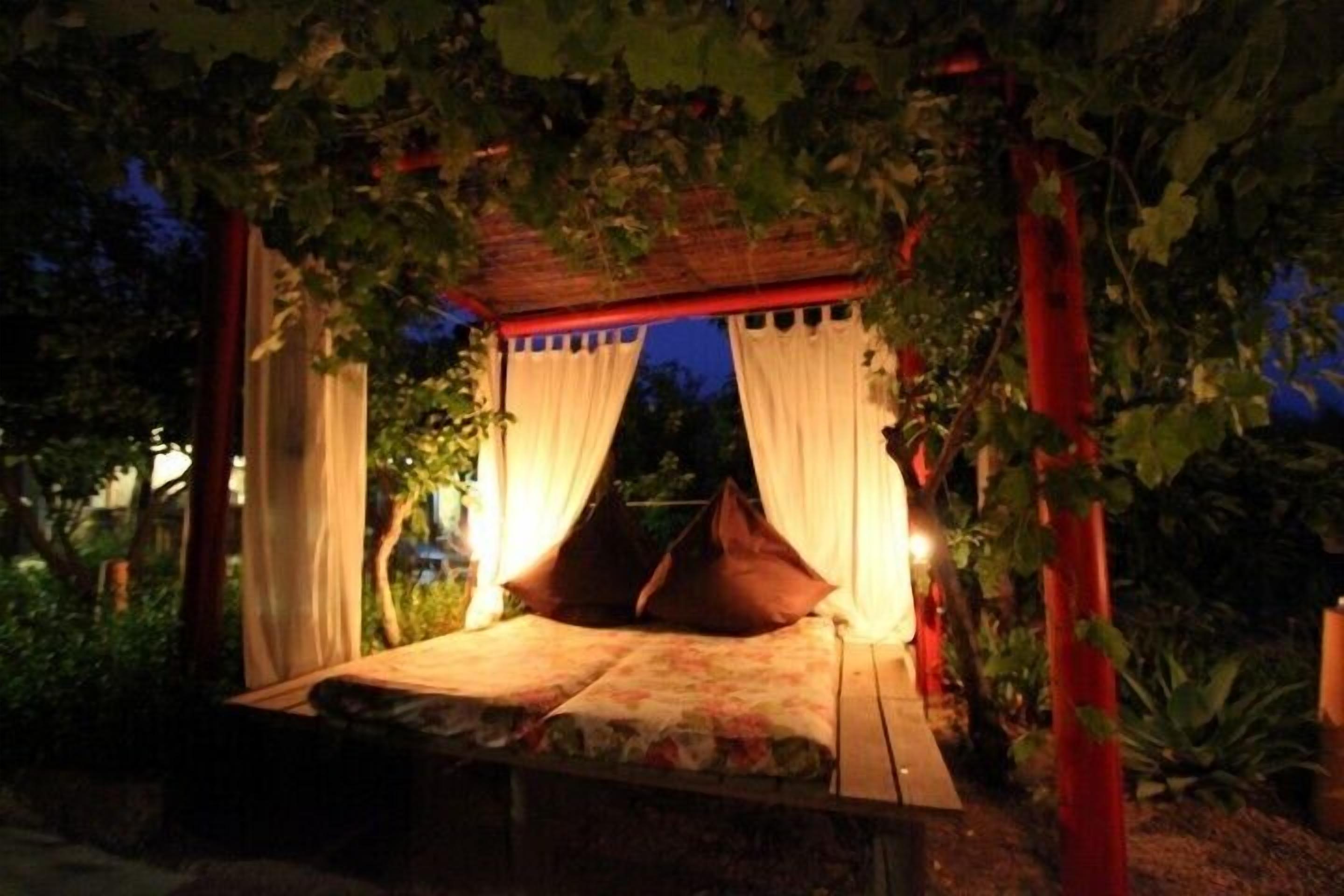 Een bed onder een pergola met gordijnen, omringd door groen.
