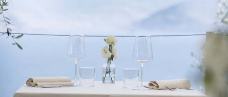 Ein Tisch ist gedeckt mit zwei Weingläsern, einer Blumenvase mit weißen Blumen und gefalteten Servietten. Der Tisch trägt eine weiße Tischdecke und bietet einen Blick auf eine Berglandschaft.