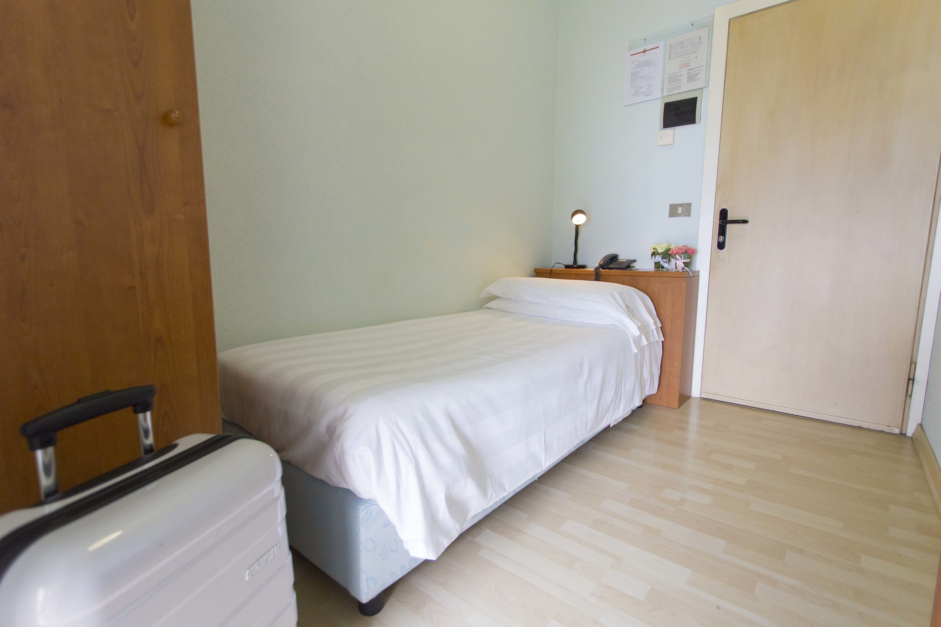 Una habitación de hotel con una cama, una mesita de noche con lámpara, una puerta de madera y una maleta.