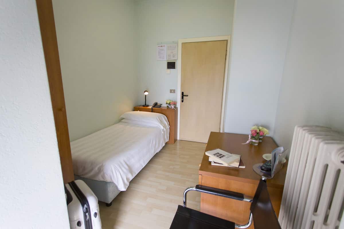A small hotel room with a bed, a desk with a lamp, a chair, a radiator, and a door with a notice on the wall.