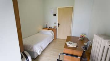 Una habitación de hotel pequeña con una cama, un escritorio con lámpara, una silla, un radiador y una puerta con un aviso en la pared.