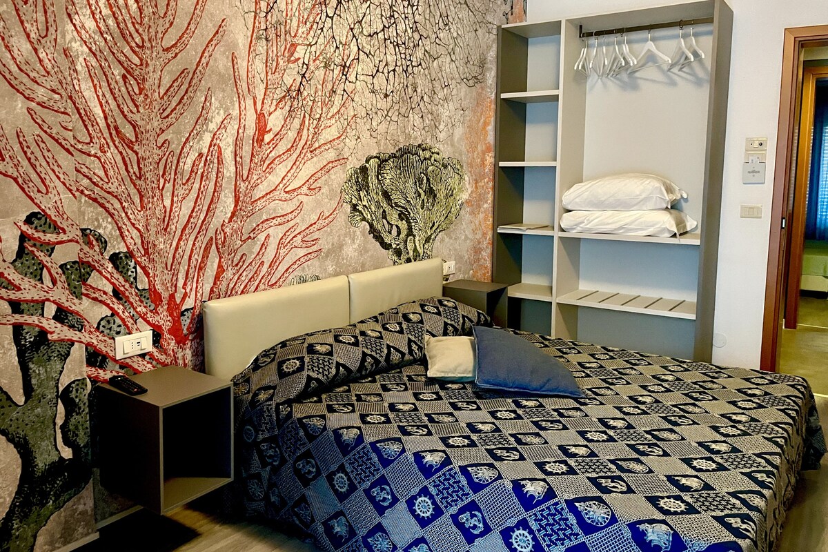 A bedroom with a patterned bedspread, a nightstand with a remote control, a wall-mounted shelf with hanging clothes, and a large abstract painting.