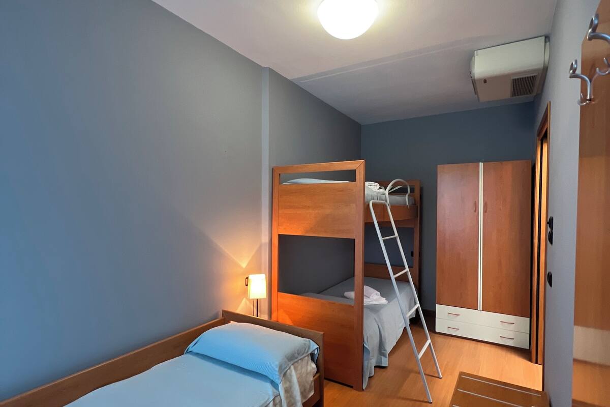Een kleine kamer met twee stapelbedden, een houten garderobe, een bureau met stoel en een klein tafeltje.