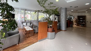 Een ruime lobby met een houten receptiebalie, grijze banken, een bijzettafeltje en potplanten.