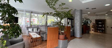 A spacious lobby with a wooden reception desk, grey sofas, a coffee table, and potted plants.