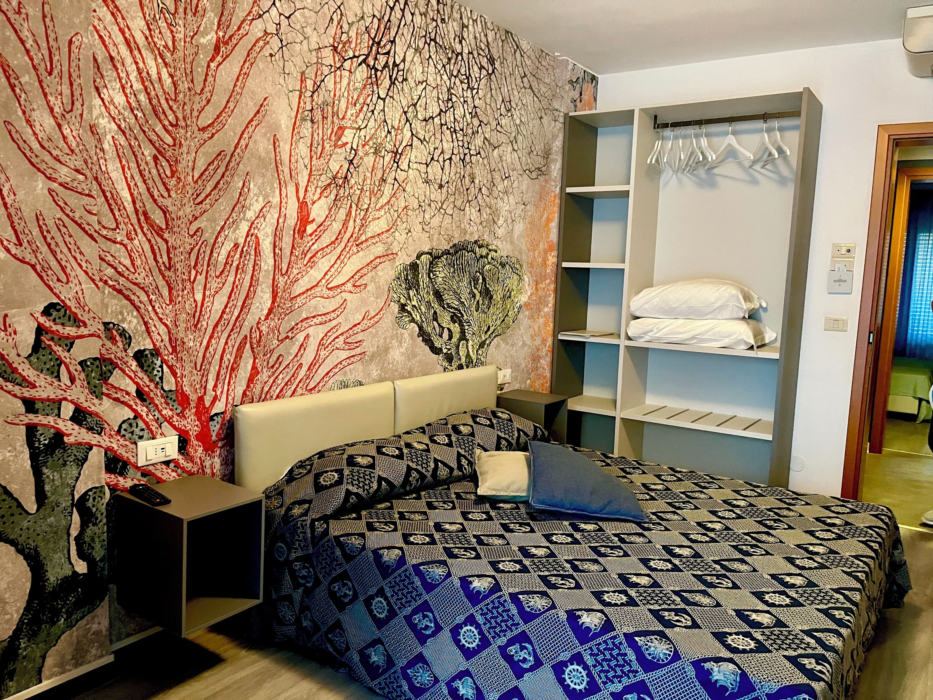 A bedroom with a patterned bedspread, a wall mural, a built-in wardrobe, and a bedside table with a remote control.