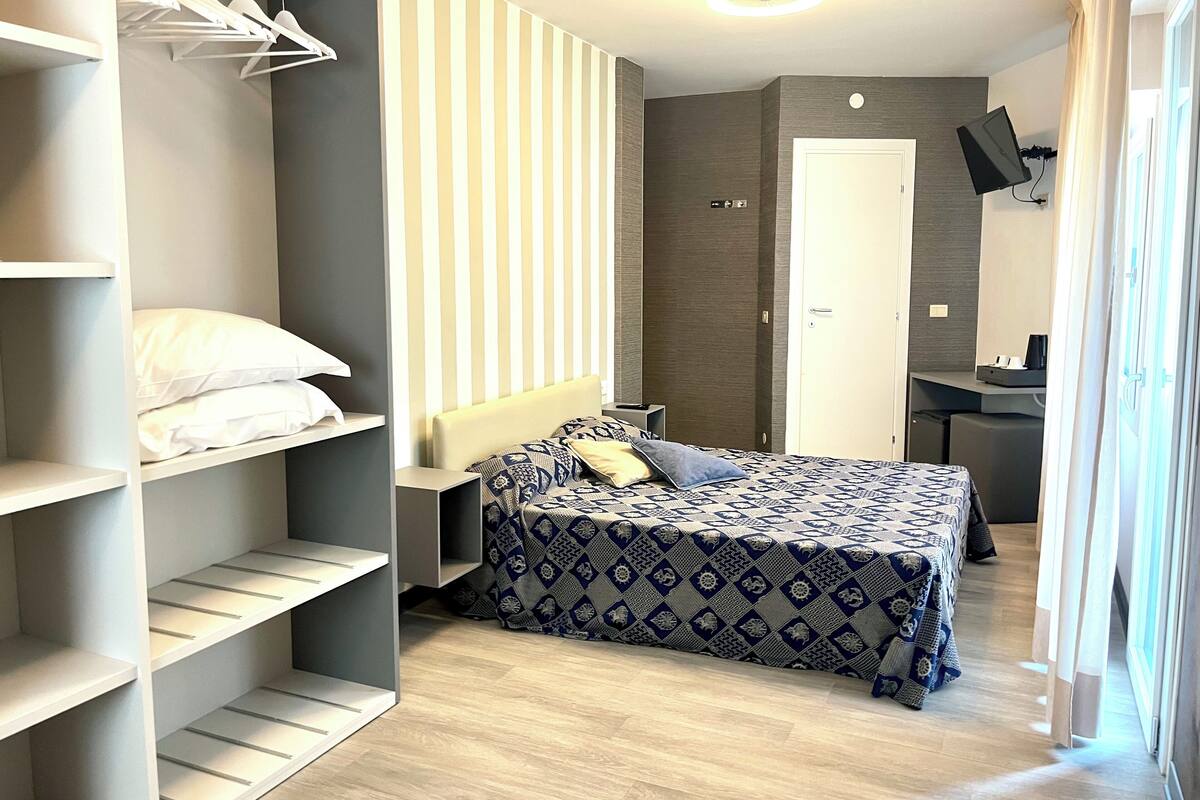 Een knus slaapkamer met een bed, een kledingkast met daarin hangende kleding, een bureau met een stoel en een aan de muur bevestigde televisie.