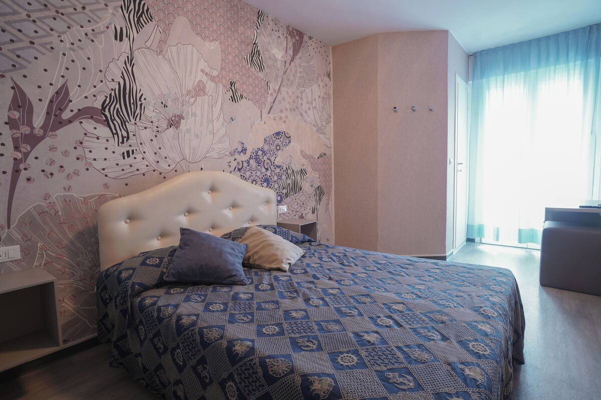 Een slaapkamer met een gestoffeerd hoofdeinde, een bed met een decoratieve spreideken en een groot raam met gordijnen.