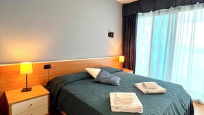 Een hotelkamer met een bed, twee bedlampen, een nachtkastje, een raam met gordijnen en een bedieningspaneel aan de muur.