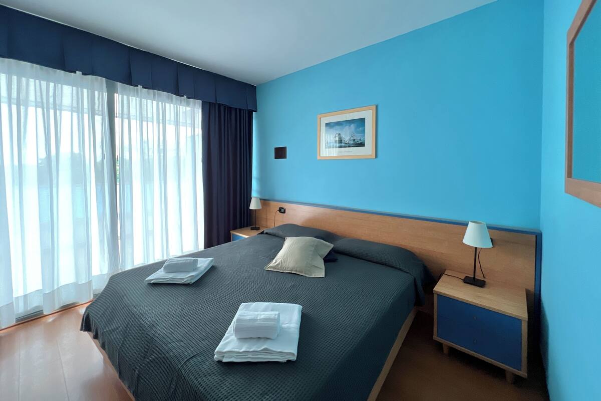 Een hotelkamer met een groot bed, twee nachtkastjes, een raam met gordijnen en een lijst met een foto aan de muur.