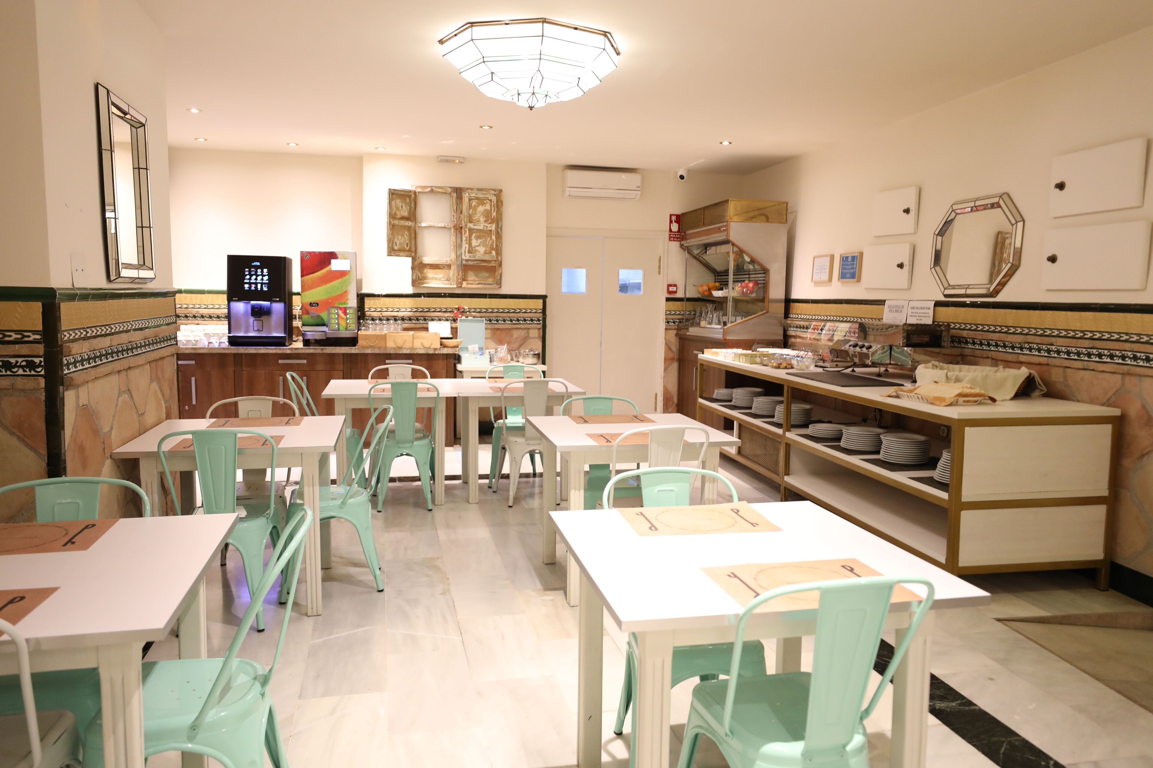 A restaurant with white tables and teal chairs, a counter with a colorful refrigerator, and a tiled backsplash.