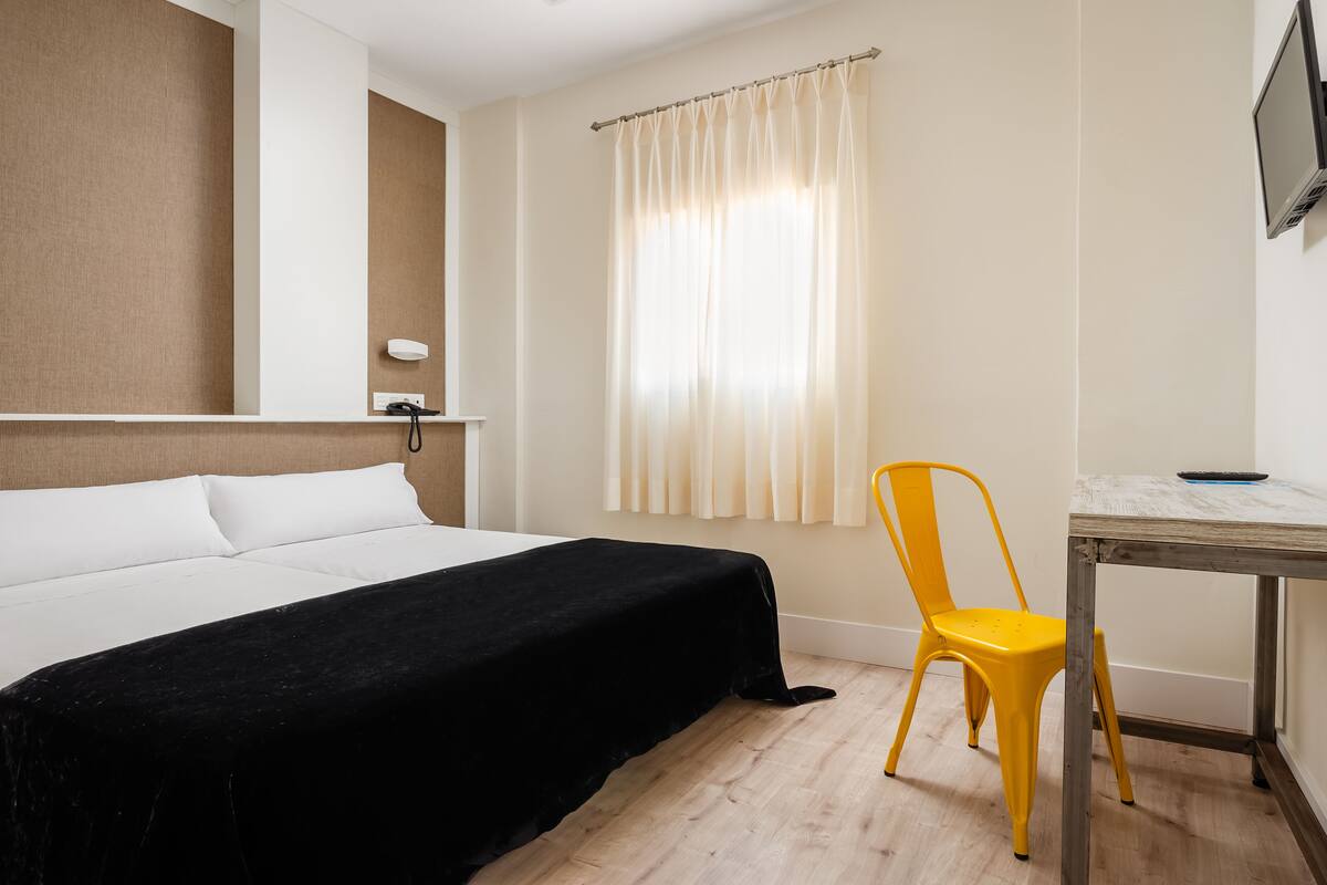 Una habitación de hotel con cama, escritorio, silla, televisor y ventana con cortinas.