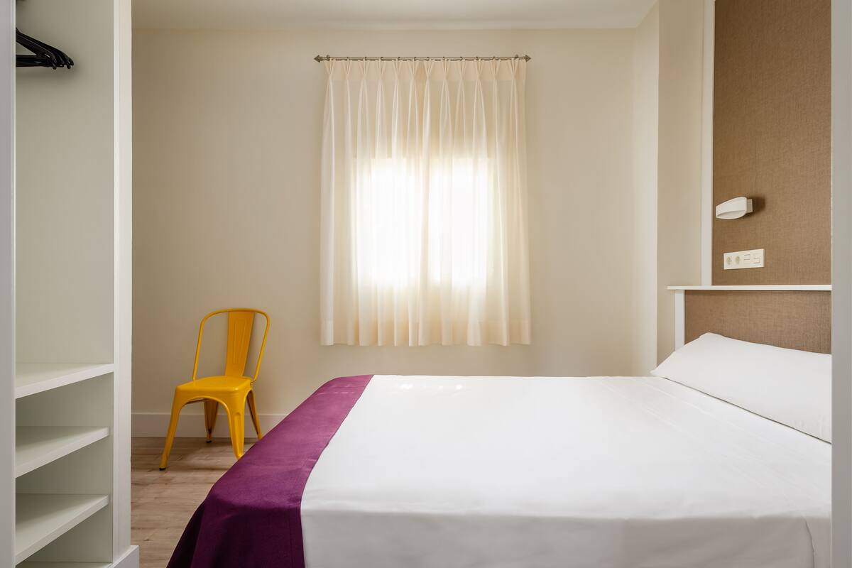 Habitación de hotel con cama, una silla, un estante y una ventana con cortinas.