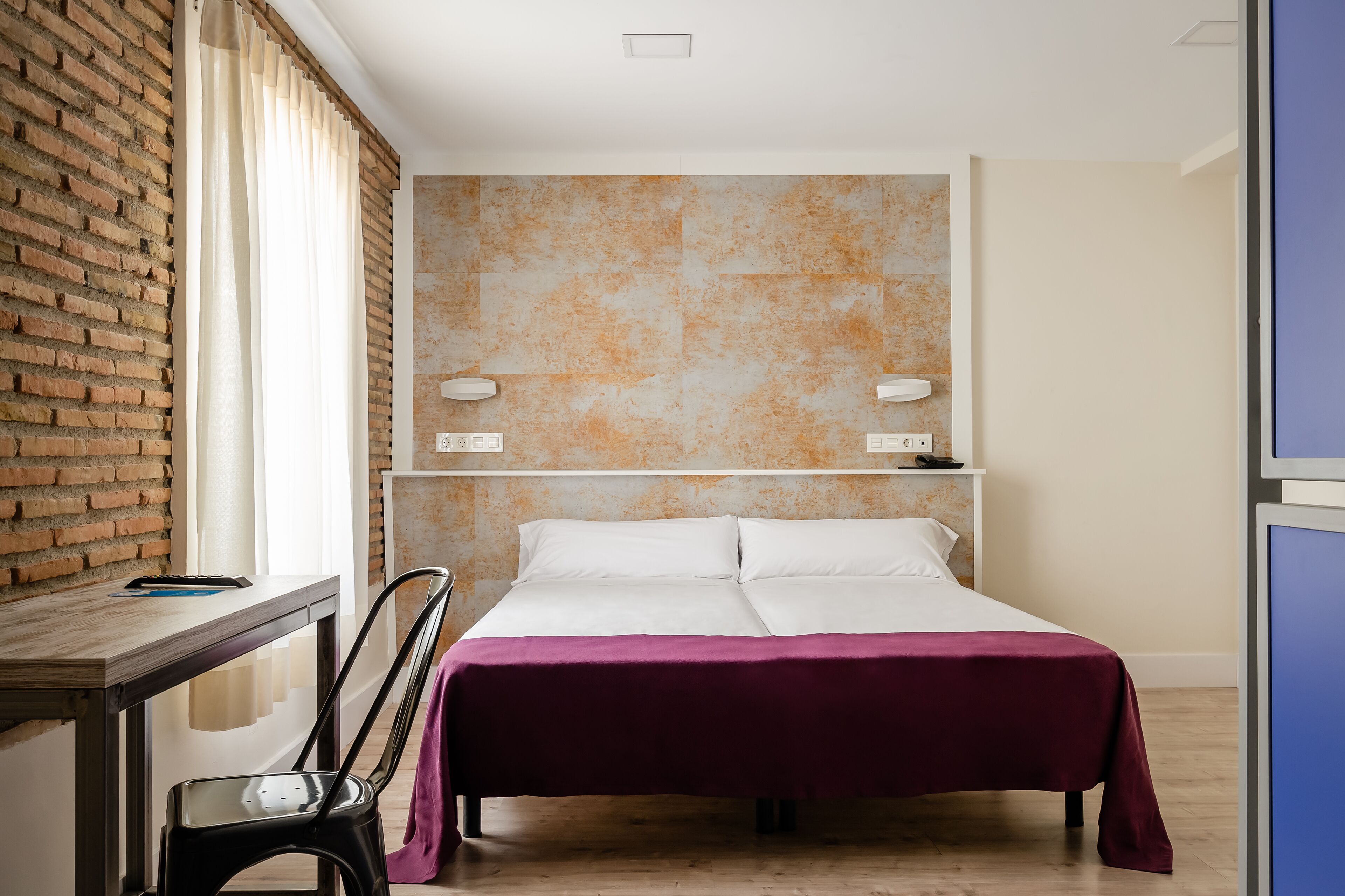 Habitación de hotel con pared de ladrillo, una cama con sábanas blancas, un escritorio con silla y una mesita de noche con lámpara.