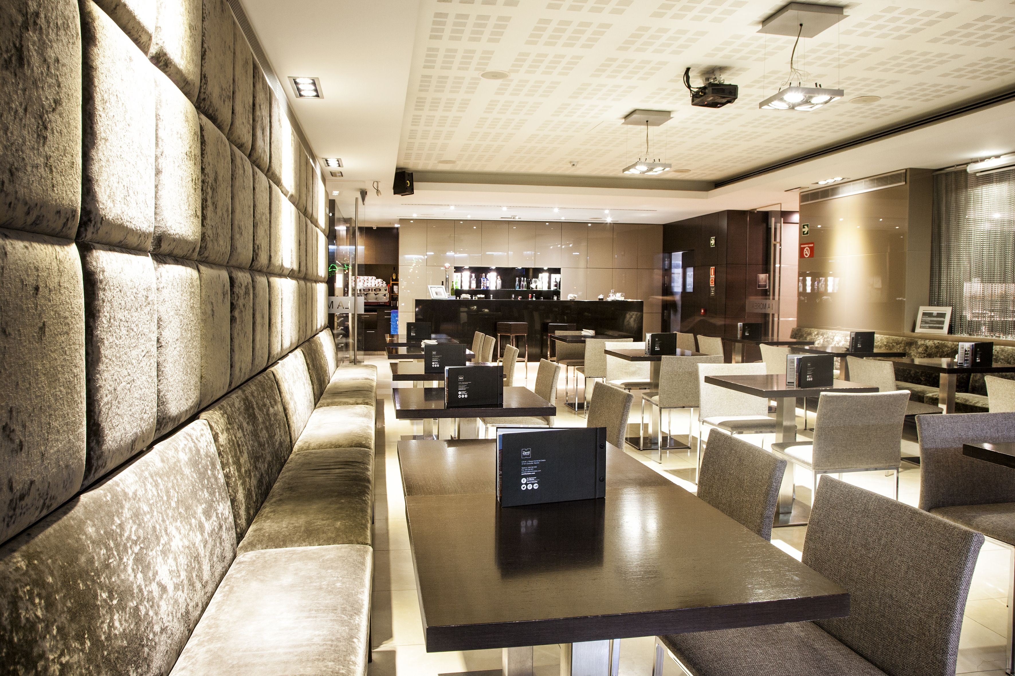 A modern restaurant with high tables and chairs, a bar area, and a seating area with cushioned benches.