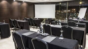 Un salón de conferencias con mesas, sillas, una pantalla de proyector y vistas a la ciudad.