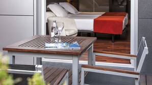 A modern hotel room with a wooden deck, a bed with a red blanket, and a table with a bottle and glass.