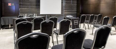 A conference room with rows of black chairs, a projector screen, and a table with water bottles.