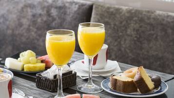 Une table mise pour le petit-déjeuner, avec une nappe sombre, des assiettes de fruits, deux verres de jus d’orange et une part de gâteau.