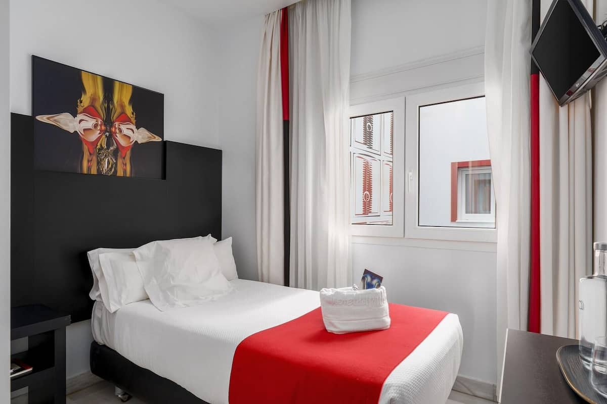 A hotel room with a bed, a TV, a painting, a window with curtains, and a small table.