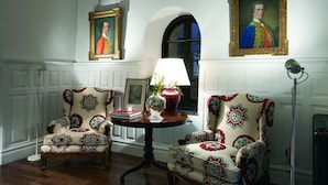 A room with two patterned armchairs, a small table with a vase and books, and two framed portraits on the wall.