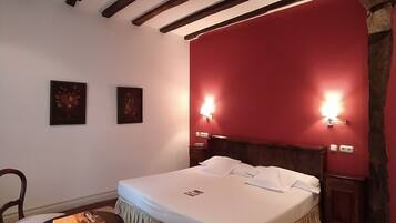 Un dormitorio con una cama grande, vigas de madera, paredes rojas, dos lámparas de noche, una silla y una mesita con libros.