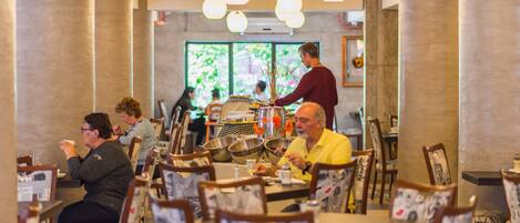 Un restaurante con mesas y sillas redondas, un camarero atendiendo a clientes y vistas a zonas verdes al exterior.