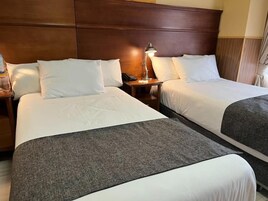 Duas camas com roupa de cama branca, cabeceira de madeira, mesa de cabeceira com abajur e manta cinza.
