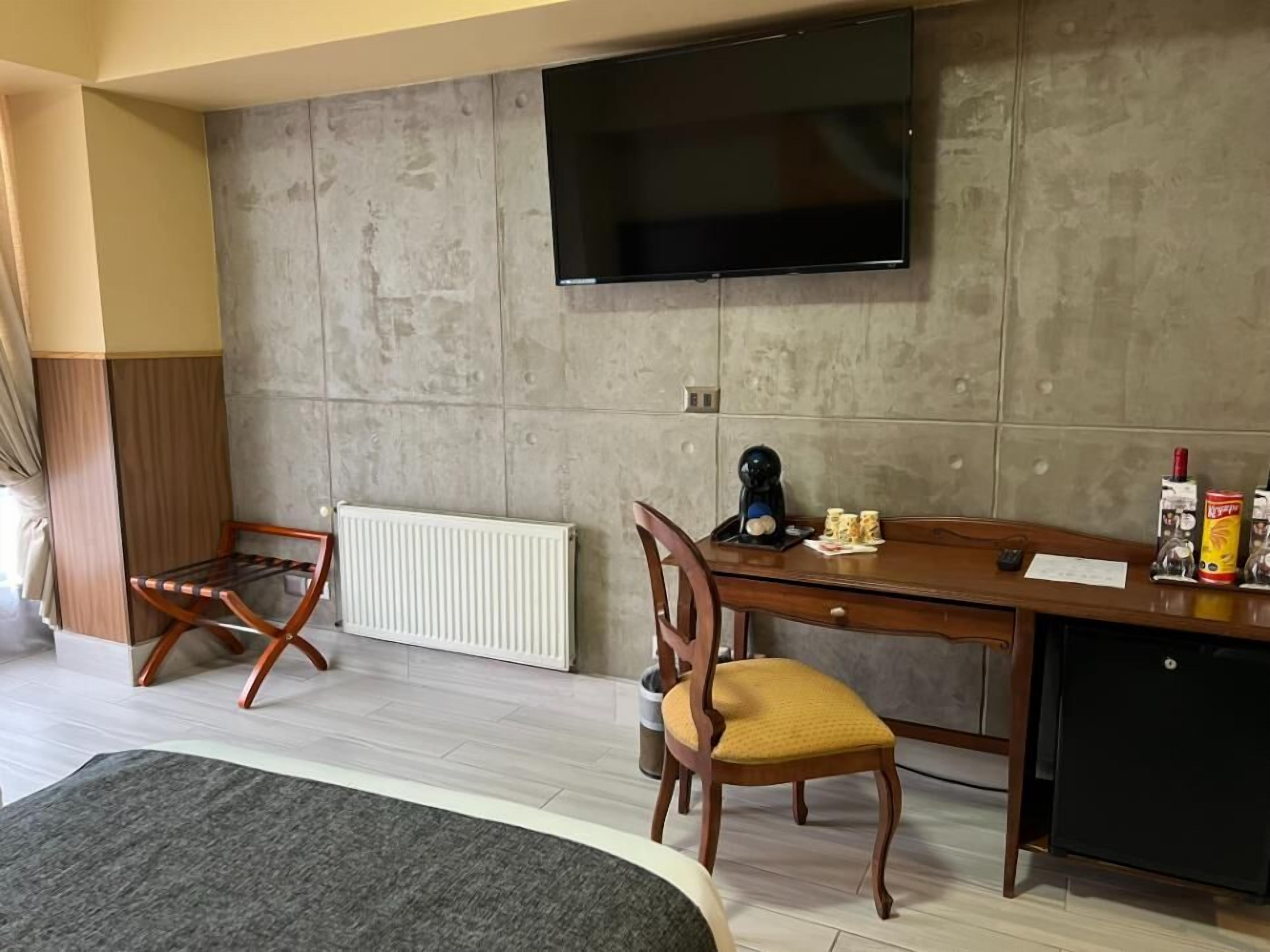 A room with a bed, a desk with a chair, a radiator, a television mounted on the wall, and a small cabinet.