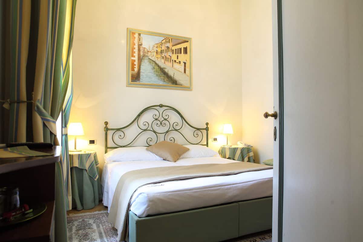 A neatly made bed with a headboard, two bedside lamps, a framed picture of a canal, and a door to the right.