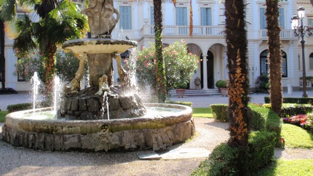 A fountain with a statue in the center, surrounded by palm trees and a building with arched windows in the background. - Villa Ducale (Dolo)