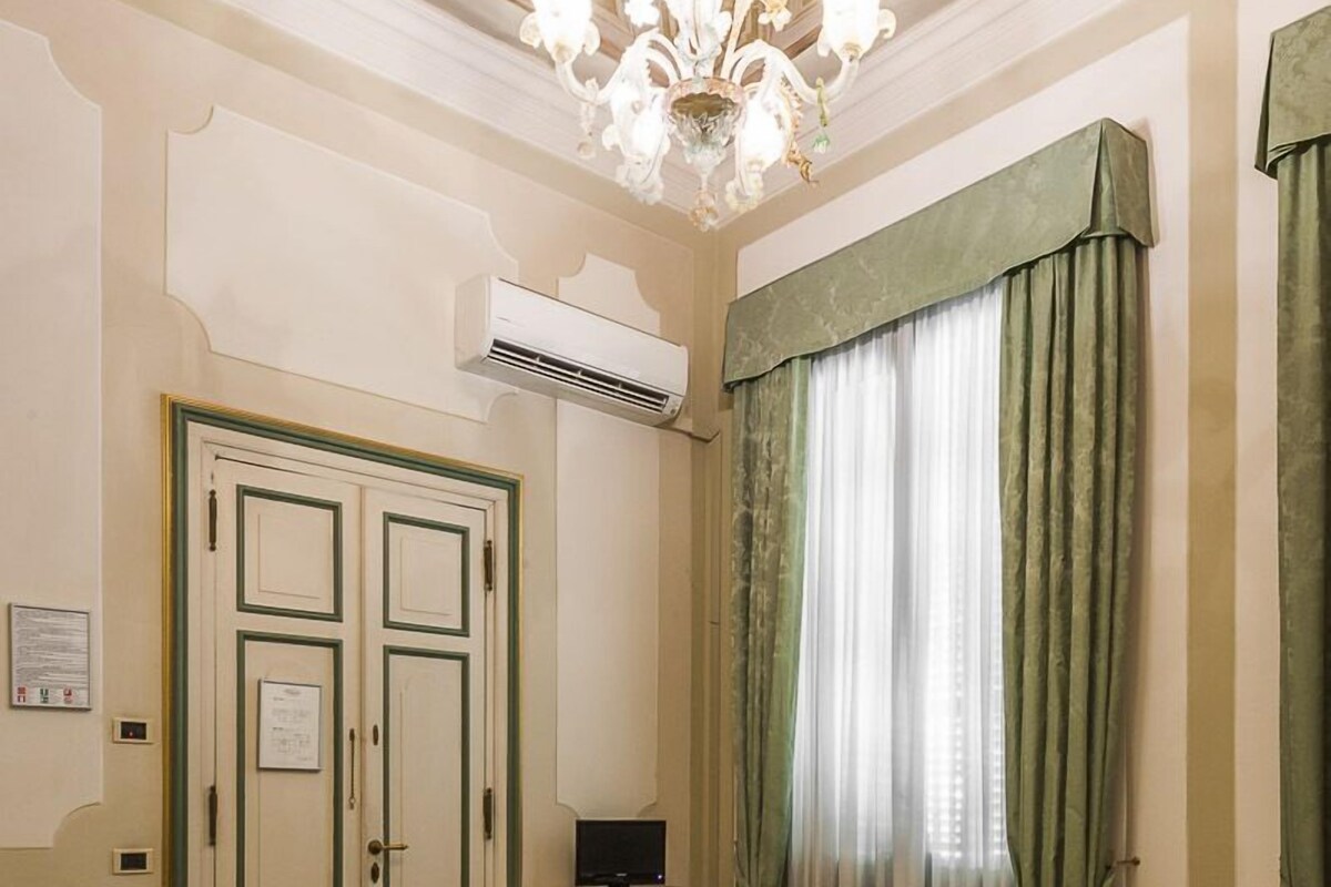 A hotel room with a bed, a desk with a computer, a chair, a small table, and a window with curtains.