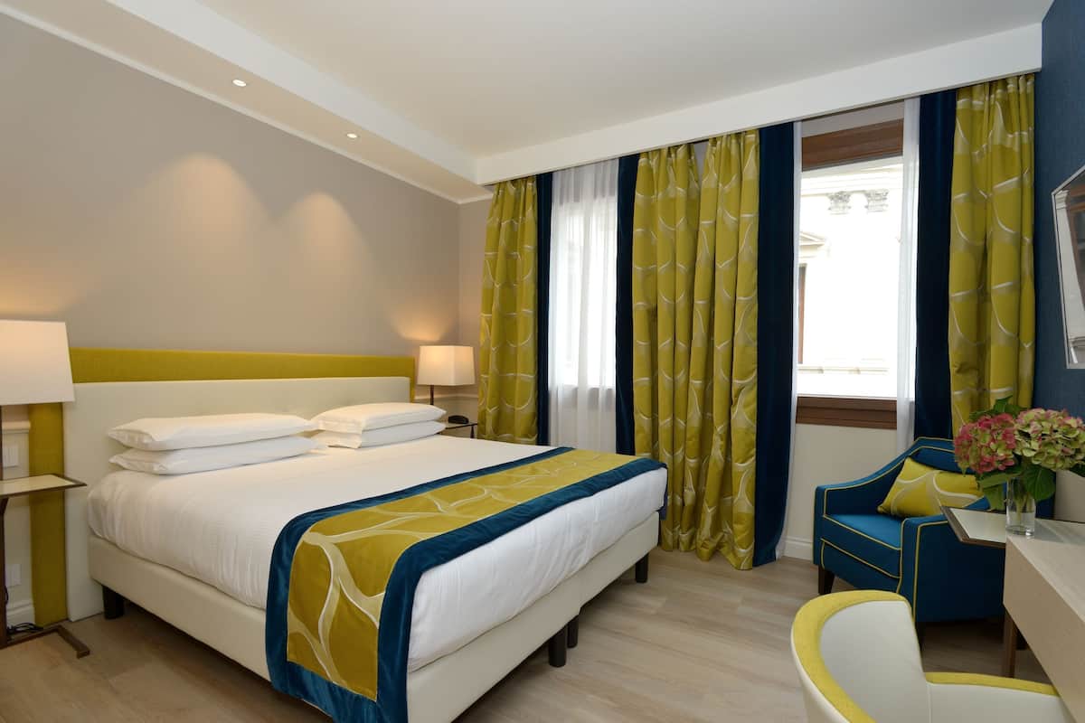 Une chambre d’hôtel avec un grand lit, deux lampes de chevet, un bureau avec une chaise et une fenêtre avec des rideaux.