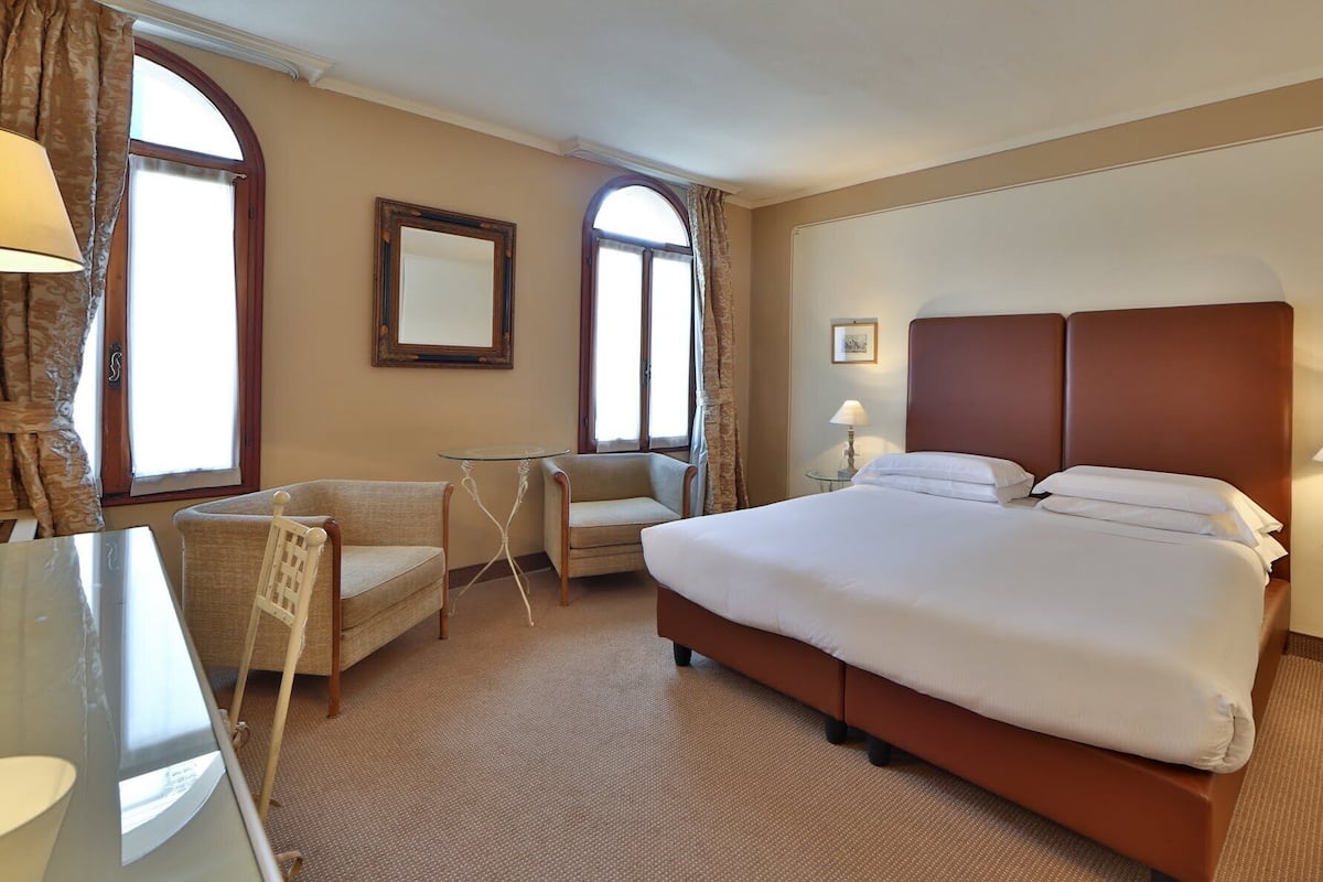 Une chambre d’hôtel avec un grand lit, deux fauteuils, une petite table et un bureau avec une lampe.