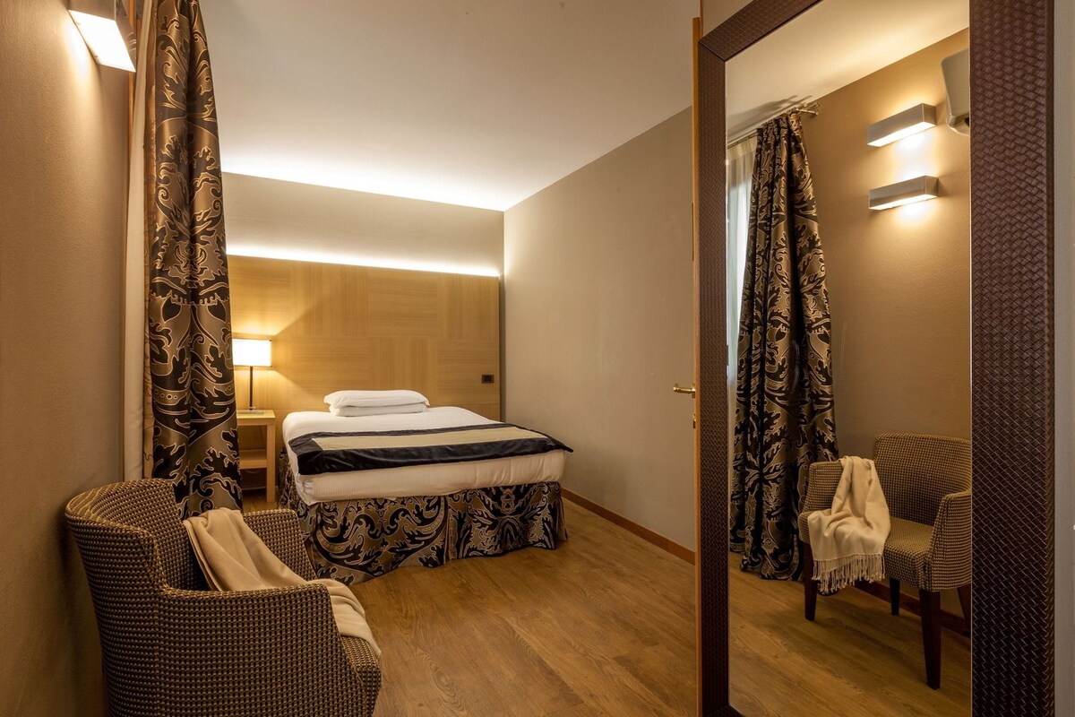 A hotel room with a bed, a chair, a bedside table, and a mirror.