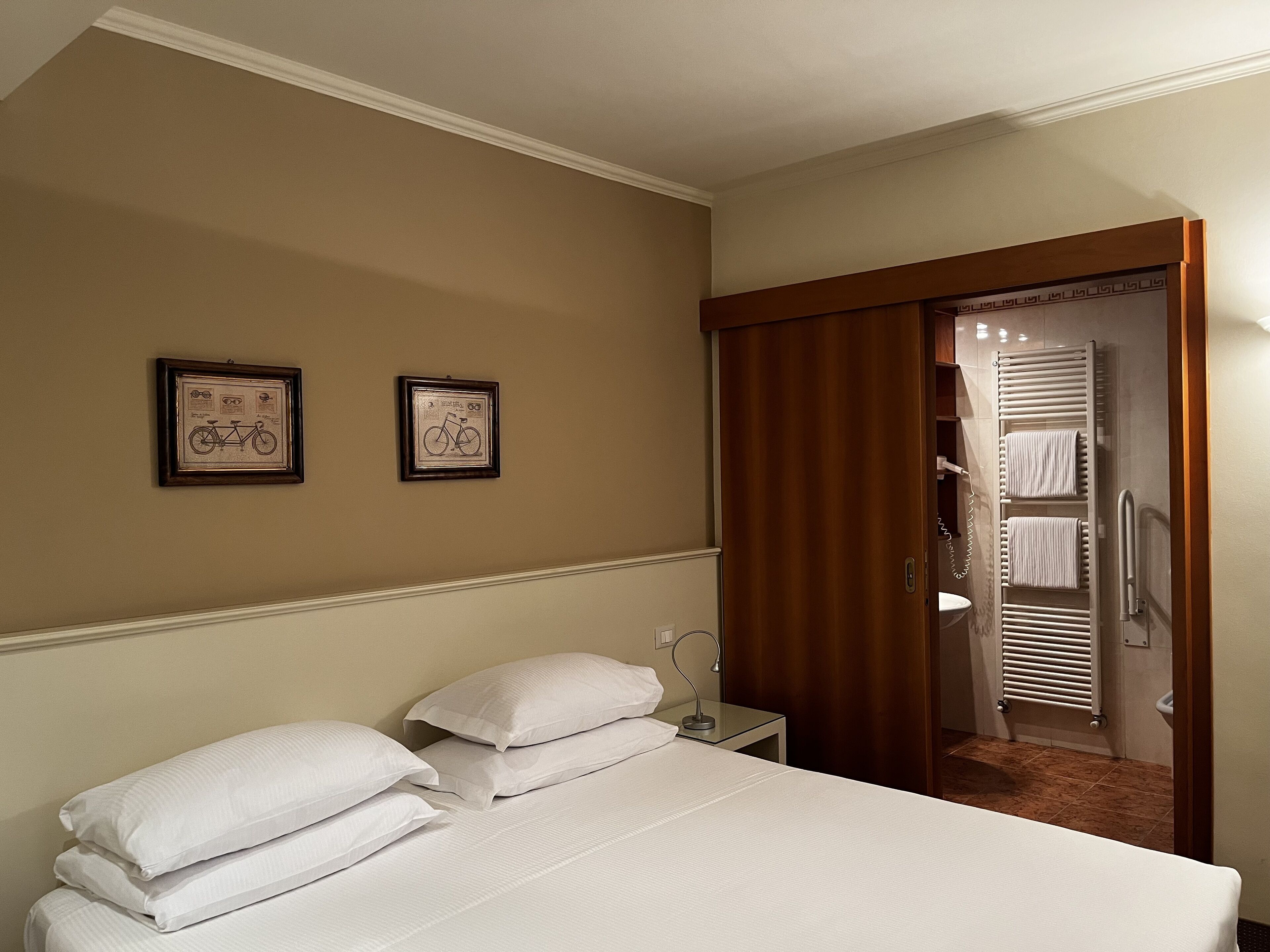 A hotel room with a bed, two framed pictures on the wall, a wooden wardrobe, and a bathroom visible through an open door.