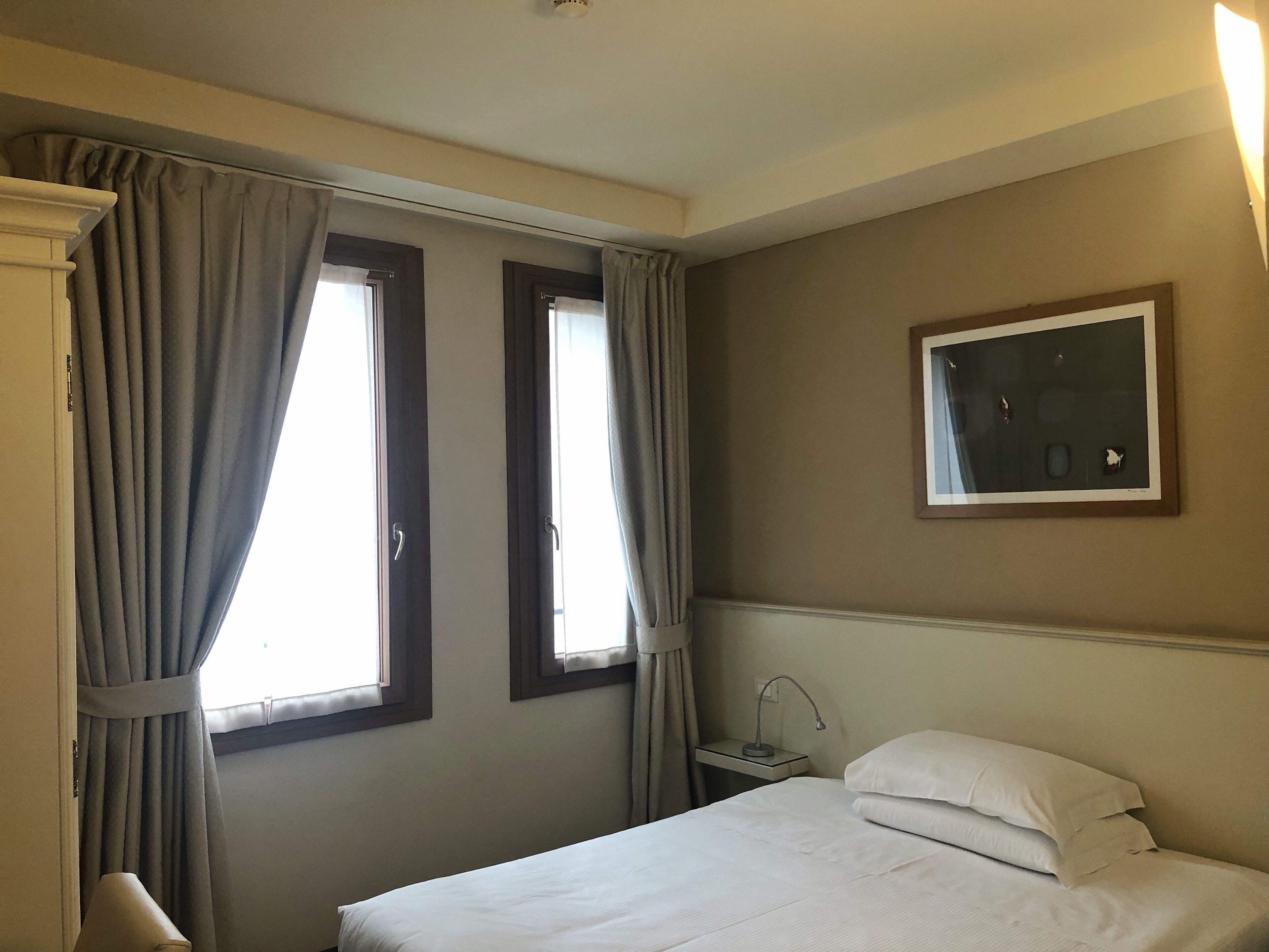 A hotel room with a bed, two windows with curtains, a bedside table with a lamp, and a framed picture on the wall.