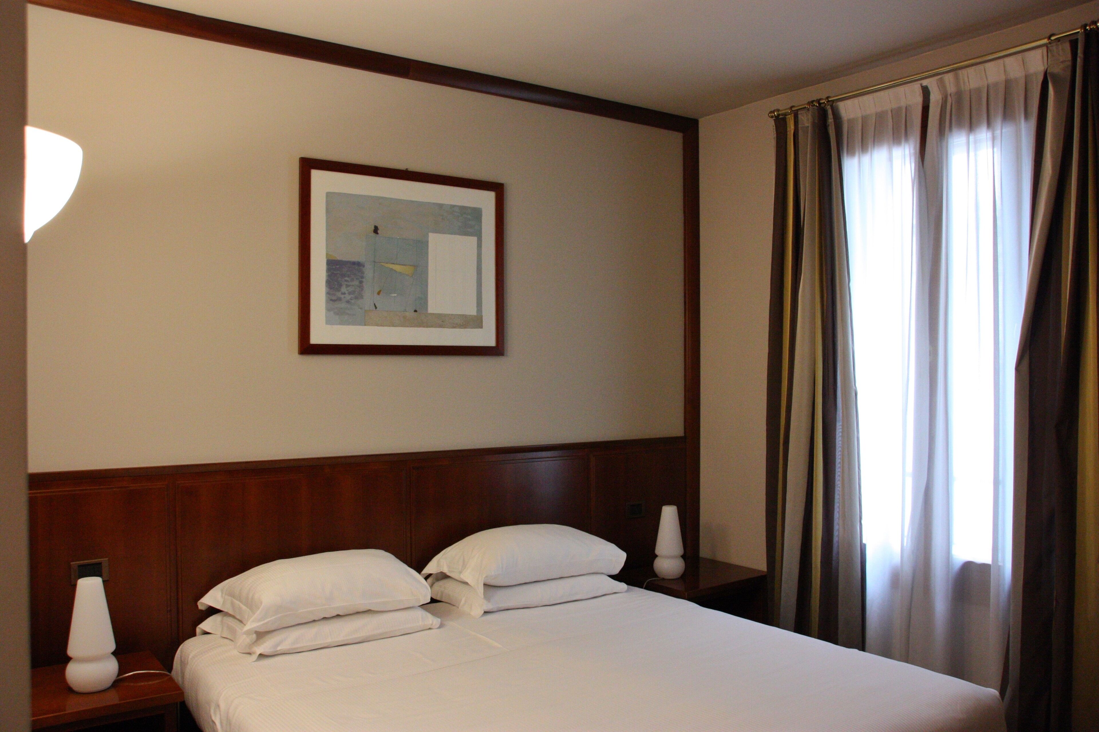 A hotel room with a neatly made bed, a framed picture on the wall, and a window with curtains.