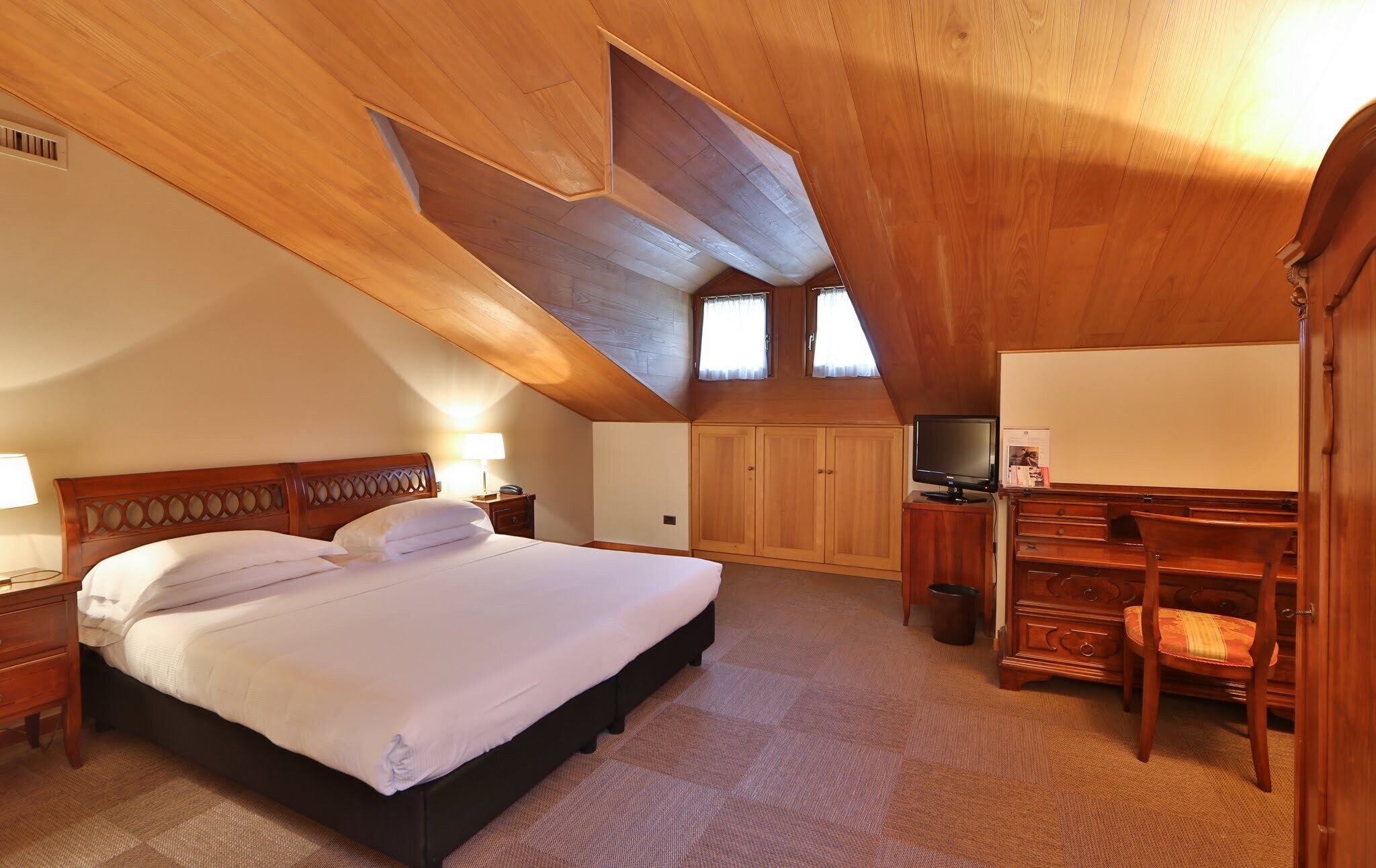 A bedroom with a large bed, wooden ceiling, built-in wardrobe, television, and a desk with a chair.