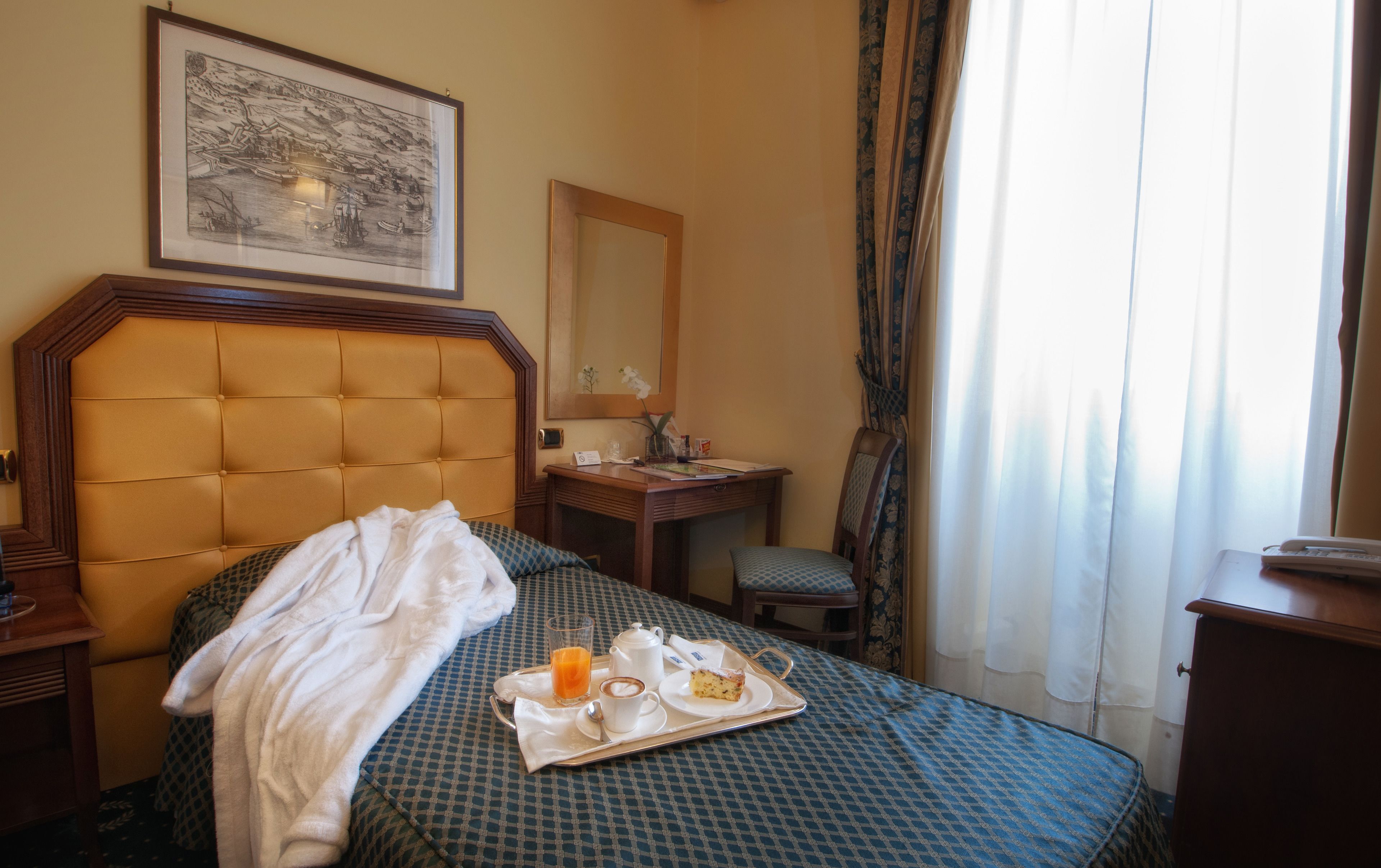 Habitació d'hotel amb un llit, una taula de nit, escriptori, cadira, finestra amb cortines i un quadre al mur.