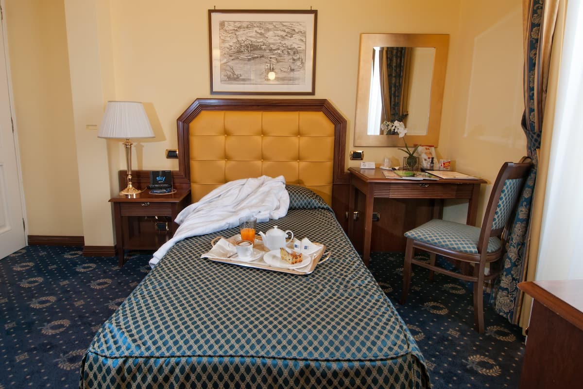 A hotel room with a large bed, a bedside table with a lamp, a chair, a desk with a mirror, and a framed picture on the wall.