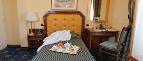 A hotel room with a large bed, a bedside table with a lamp, a chair, a desk with a mirror, and a framed picture on the wall.