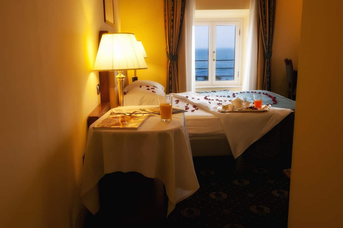 A hotel room with a bed, a bedside table with a lamp, a window with curtains, and a view of the sea.