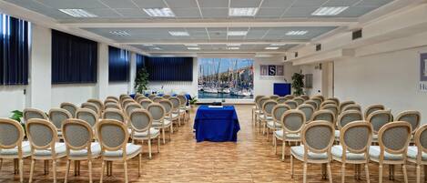 Ein Konferenzraum mit Reihen aus Holzstühlen, einem blauen Tisch im vorderen Bereich und Blick auf Boote durch große Fenster.