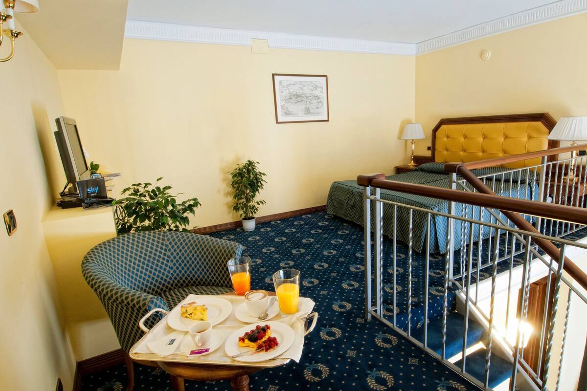 A hotel room with a dining area, a bed with a headboard, a staircase, and a potted plant.