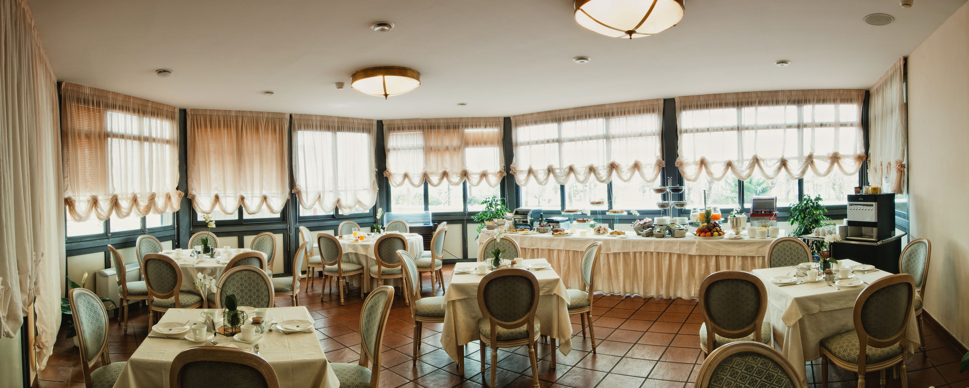 A dining area with multiple tables and chairs, large windows with curtains, and a kitchen area visible in the background.