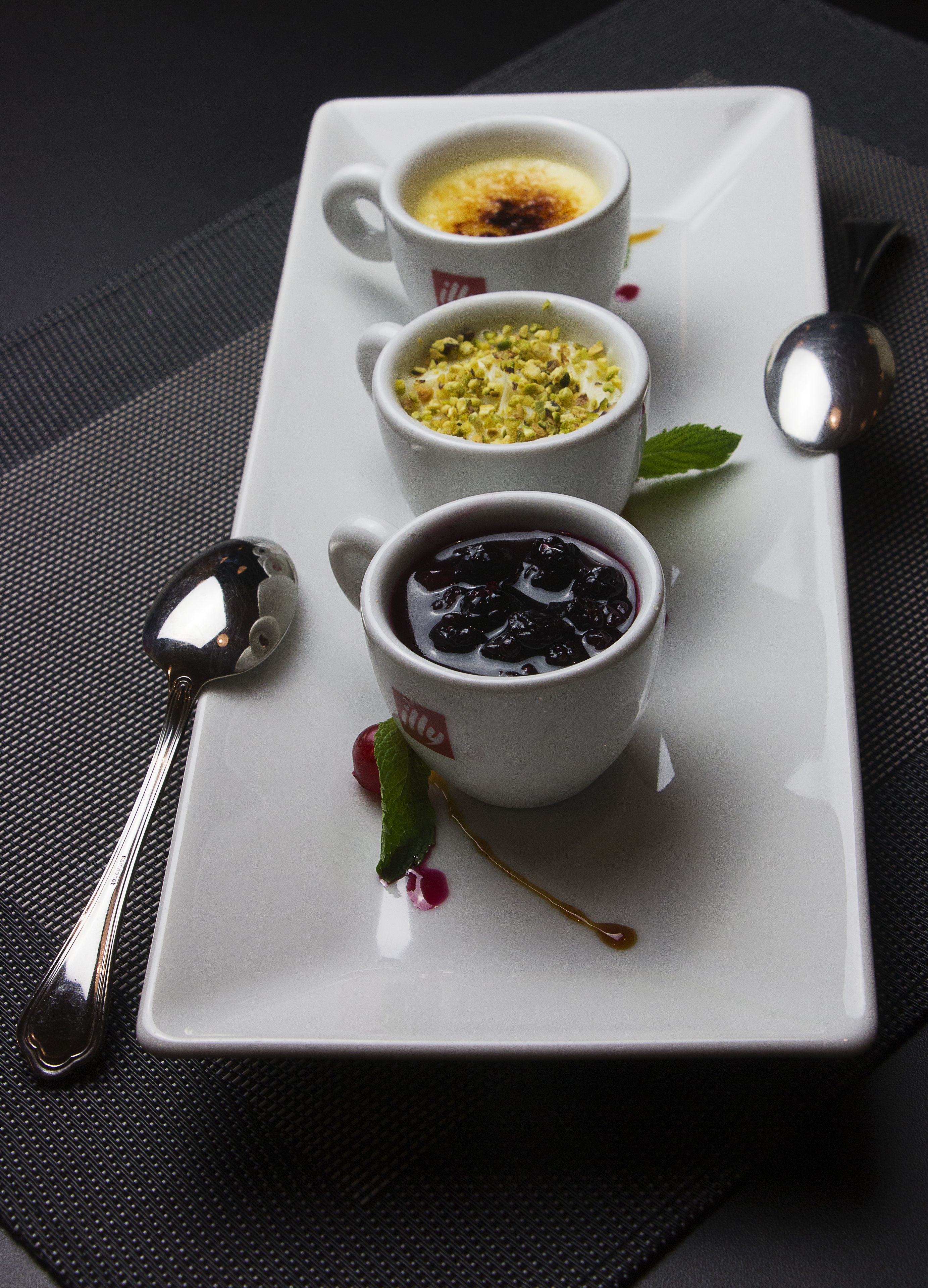 Three white cups with different desserts on a white rectangular plate, a spoon, and a small mint leaf.