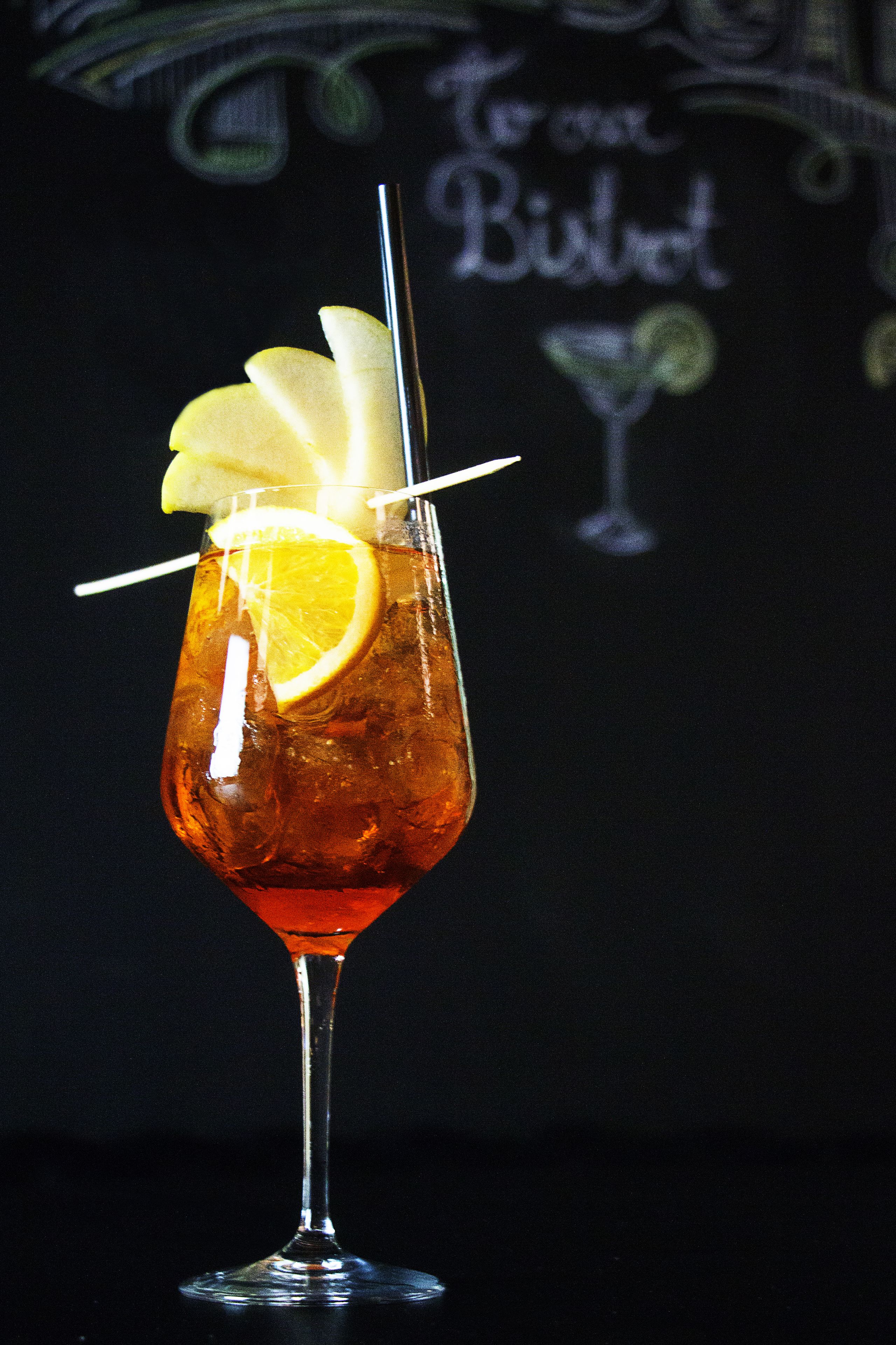 A cocktail glass with a lemon slice and a straw, set against a dark background with a chalkboard displaying "To our Bistrot".