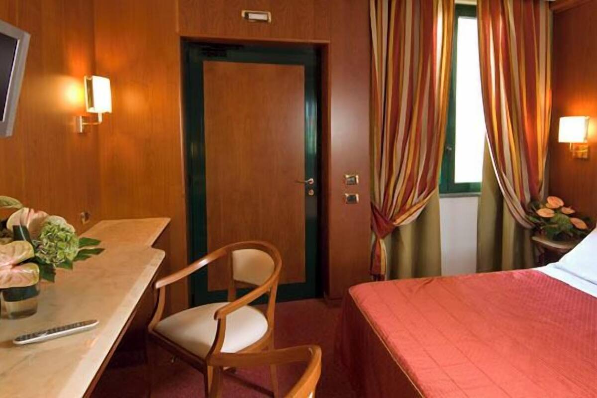 Habitación de hotel con cama, escritorio, silla, televisor y ventana con cortinas.